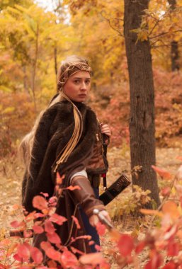 Sonbahar ormanında kılıçlı savaşçı kızlar. Savaşçı kadın kıyafeti giymiş, kahverengi kürklü, ortaçağ elbiseli, savaşçı kız çelik kılıcı elinde tutuyor.