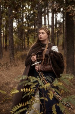 Sonbahar ormanında kılıçlı savaşçı kızlar. Kahverengi kürk pelerinli, ortaçağ elbisesi giymiş, tarihi bir kadın gibi giyinmiş bir sarışın. Savaşçı kız çelik kılıcı elinde tutuyor.. 