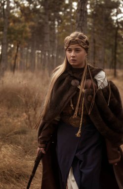 Tarihi savaşçı kostümlü sarışın kızın yakın çekimi. Uzun kürk kahverengi bir ceket, eski moda bir elbise. Ellerinde antika tabanca var. Ormanda hızla yürür..