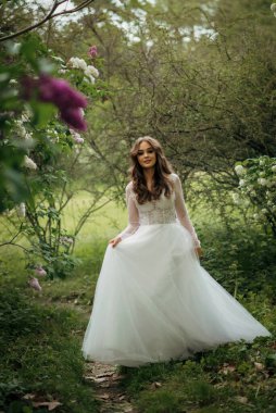 Parkta zarif gelinlik içinde genç ve güzel bir kız gelin. Baharda çiçek açan ağaçlar. Dantel ve şifondan yapılmış uzun bir elbise. Kız gülümsüyor. Avrupa düğün modası konsepti.