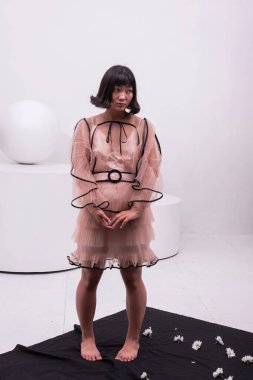 Kısa saçlı Asyalı bir kızın stüdyo fotoğrafı. Onun pembe allık ve soluk pembe bir elbisesi olan yumuşak bir makyajı var. Beyaz kasımpatı olan siyah bir kumaşın üzerinde yalınayak duruyor..