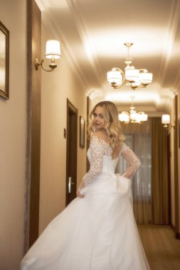 Otel koridorunda poz veren uzun beyaz elbiseli güzel sarışın gelin. Korseli ve omuzları açık şık bir elbise. Sarı saçlar bol ve şık bir stili var. Gülümsüyor ve dans ediyor.. 