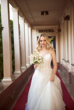 Güzel sarışın gelin elinde bir buketle otelin açık galerisinde yürüyor. Omuzları açık, uzun bir gelinlik. Dalgalı sarı saçlar, açık omuzlar. Gülümsüyor ve kameraya bakıyor..