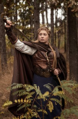 Elinde kılıç olan savaşçı bir kızın sanat fotoğrafı. Savaşçı bir kadın kostümü giymiş, pelerinli ve klasik elbiseli bir sarışın. Savaşçı kız kılıcını kaldırdı. Kameraya bak..