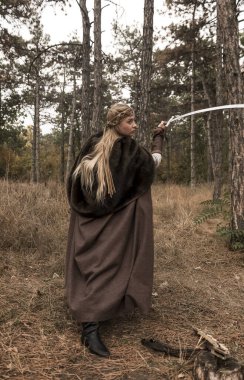 Ormanda elinde kılıç olan savaşçı kız. Savaşçı kadın kostümlü, uzun pelerinli bir sarışın. Savaşçı kız, kılıcını kaldırdı, düşmana saldırdı, kameraya baktı..