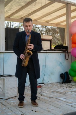 Odessa, Ukrayna - 05 Nisan 2025: Palto giyen bir adam saksafon çalar, beyaz ahşaptan yapılmış geçici bir sahnede ve renkli balonlarla.