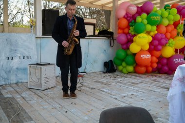 Odessa, Ukrayna - 05 Nisan 2025: Palto giyen bir adam saksafon çalar, beyaz ahşaptan yapılmış geçici bir sahnede ve renkli balonlarla