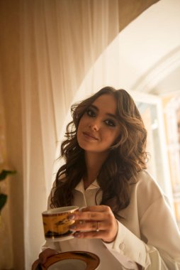 Genç, romantik esmer, uzun dalgalı saçlı, elinde fincan ve fincan tabağıyla bir odada. Beyaz ipek bir takım elbise giymişti. Hafif bir gülümsemeyle kameraya bakar. yakın plan