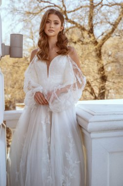 Geleneksel uzun beyaz elbiseli esmer gelin baharda otel balkonunda poz veriyor. Uzun kollu, uzun beyaz elbise, korse ve açık omuzlar. Gelin portresi, düğün makyajı, şık gelin