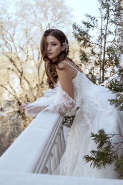 Geleneksel uzun beyaz elbiseli esmer gelin baharda otel balkonunda poz veriyor. Uzun kollu beyaz elbise, korse ve açık omuzlu. Klasik mimarisi olan bir otel. moda gelin