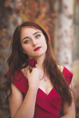 Ellerinde bir gülle sonbahar ağaçlarının arka planında duran kırmızı elbiseli genç bir kızın portresi. Uzun siyah saçlı düşünceli ve romantik bir kız.. 