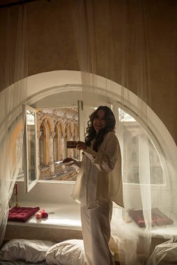 Uzun dalgalı saçlı genç esmer bir fincan kahve ve kahkahalarla pencerenin önünde duruyor. Beyaz ipek bir takım elbise giymişti. Yatağın yanında duruyor. Beyaz perdeli kemerli pencere.