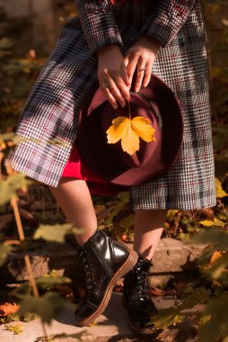 Siyah botları, paltosu, elinde şapkası ve sarı yaprağı olan bir kız parkta duruyor. Sonbahar konsepti, sonbahar modası. Botlar yakın plan, yüz yok.