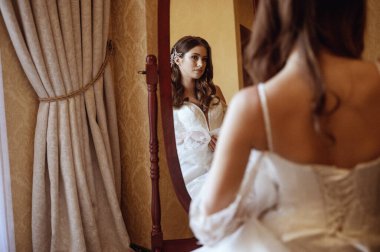 Uzun geleneksel beyaz elbiseli gelin düğünden önce otel odasında aynanın önünde duruyor. Onun uzun saçı beyaz çiçeklerle süslenmiş. Arka plan, bağcıklı beyaz korse. Ön plan bulanık