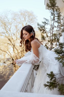 Geleneksel uzun beyaz elbiseli esmer gelin baharda otel balkonunda poz veriyor. Uzun kollu beyaz elbise, korse ve açık omuzlu. Klasik mimarisi olan bir otel. düğün makyajı