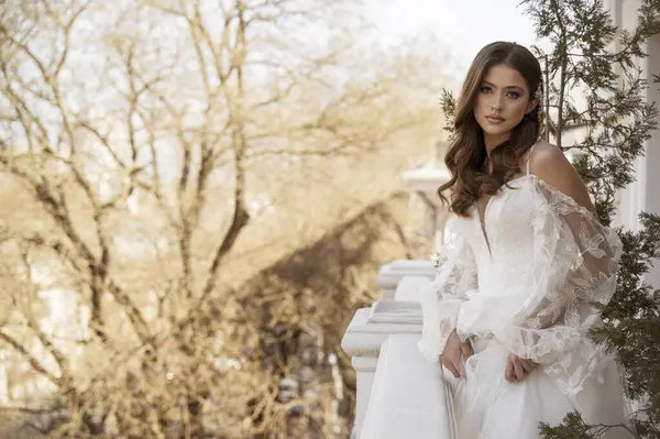 Geleneksel uzun elbiseli esmer gelin baharın başında beyaz otel balkonunda poz veriyor. Uzun kollu beyaz elbise, korse, açık omuzlar. düğün makyajı, düğün saç stili.