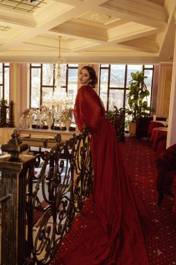 Lüks otel odasının arka planına karşı kırmızı uzun elbiseli bir model. Otel lobisinde demir parmaklıklı, uzun gece elbisesi giymiş zarif genç bir bayan. Akşam makyajı ve saç stiliyle..