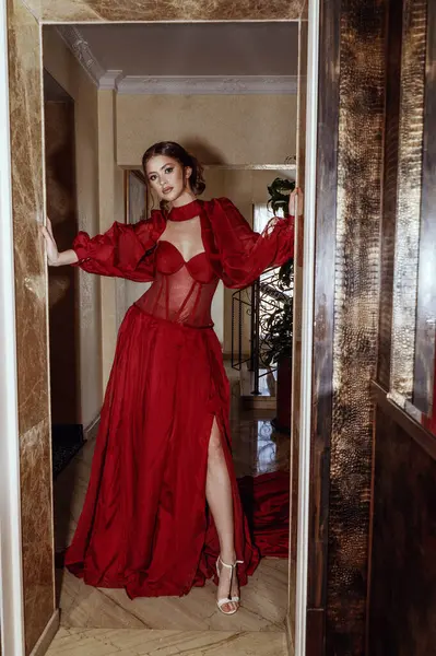 Kırmızı elbiseli, akşam makyajlı ve saç stili zarif genç bir bayan. Asansörün yanında duran, kameraya bakan, uzun kollu korseli bir elbise giyen, otelin içinde çekim yapan bir kız..