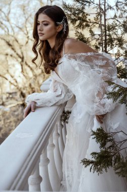 Geleneksel uzun elbiseli esmer gelin baharda beyaz otel balkonunda poz veriyor. Uzun kollu beyaz elbise, korse ve açık omuzlu. Gelin portresi, düğün makyajı, şık gelin