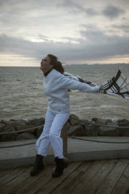 Beyaz eşofmanlı güzel genç kız fırtınalı havada deniz kıyısında duruyor. Onun sıcak büyük bir eşarbı var. Kafesteki atkı, hafif. Dramatik bir fotoğraf. Saçları ve atkısı rüzgârla savruluyor.