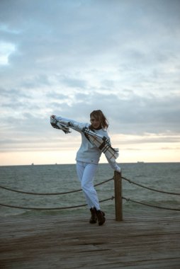Beyaz eşofmanlı güzel genç kız fırtınalı havada deniz kıyısında duruyor. Onun sıcak büyük bir eşarbı var. Kafesteki atkı, hafif. Dramatik bir fotoğraf. Saçları ve atkısı rüzgârla savruluyor.