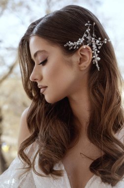 Geleneksel beyaz elbiseli esmer gelin profili yakın plan. Dalgalar halinde uzun saçlar, kenarlarında küçük inciler olan beyaz süslemeler. Gelin portresi, düğün makyajı ve saç stili.