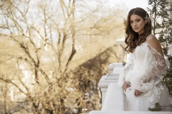 Geleneksel uzun elbiseli esmer gelin baharın başında beyaz otel balkonunda poz veriyor. Uzun kollu beyaz elbise, korse, açık omuzlar. Gelinin portresi, düğün makyajı, saç stili..
