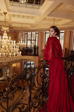 Lüks bir otel odasının arka planında kırmızı, uzun bir elbise giymiş bir model. İçinde kristal avizeler, aynalar ve içeride sahte elementler var. Otel lobisinde zarif genç bir bayan..