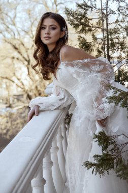 Geleneksel uzun elbiseli esmer gelin baharda beyaz otel balkonunda poz veriyor. Uzun kollu beyaz elbise, korse ve açık omuzlu. Gelin portresi, düğün makyajı, şık gelin.