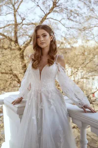 Geleneksel uzun beyaz elbiseli esmer gelin baharda otel balkonunda poz veriyor. Uzun kollu, uzun beyaz elbise, korse ve açık omuzlar. Gelin portresi, düğün makyajı