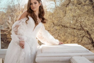 Geleneksel uzun elbiseli esmer gelin baharın başında beyaz otel balkonunda poz veriyor. Uzun kollu beyaz elbise, korse, açık omuzlar. düğün makyajı, saç stili.