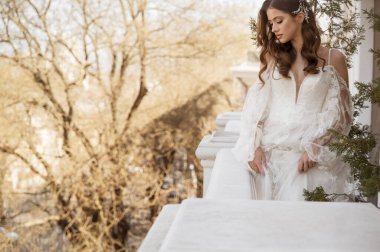 Geleneksel uzun elbiseli esmer gelin baharda beyaz otel balkonunda poz veriyor. Uzun kollu beyaz elbise, korse, açık omuzlar. Gelin portresi, düğün makyajı, şık gelin.