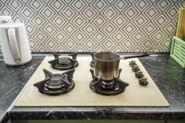 Mutfağın içindeki gaz sobasının üstündeki tencereye yakın çekim. Açık bej gaz paneli, yakınlarda beyaz elektrikli su ısıtıcısı..