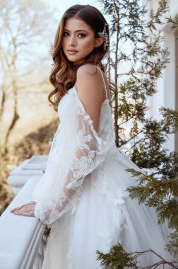 Geleneksel uzun elbiseli esmer gelin baharda beyaz otel balkonunda poz veriyor. Uzun kollu beyaz elbise, korse ve açık omuzlu. Düğün makyajı, moda gelin.