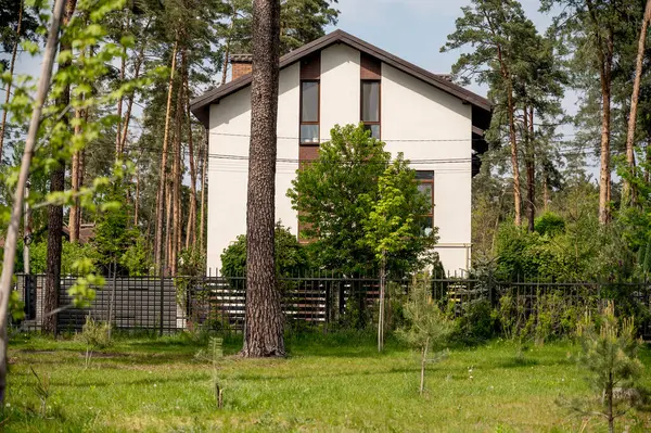 Ön cephesi ve iki katlı kahverengi çatısı olan yeni evin güzel dış görünüşü. Köyde çam ormanlarıyla çevrili beyaz evler. Evin etrafında çeşitli ağaçlar var.. 