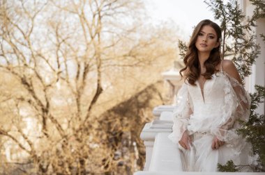 Geleneksel uzun elbiseli esmer gelin baharda beyaz otel balkonunda poz veriyor. Uzun kollu beyaz elbise, korse, açık omuzlar. Gelinin portresi, düğün makyajı, düğün saç stili..