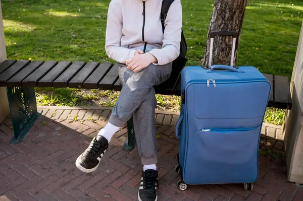 Parkta elinde telefon ve mavi bir bavulla oturan bir turist. Telefonu ellerinin arasında tutuyor ve ona bakıyor. Açık bej kazaklı ve gri pantolonlu. Avrupa 'da Turizm