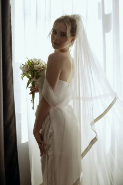 Uzun peçeli sarışın gelin elinde zambak buketiyle pencerenin yanında uzun beyaz bir elbise içinde duruyor. Minimalist iç mekan, otel ya da ev. Yan tarafında kesik olan uzun elbiseli gelin.