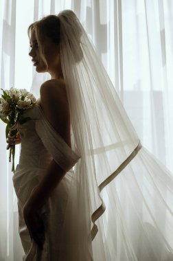 Sarisin gelinin silueti, duvagli, uzun beyaz bir elbiseyle, pencerenin yaninda bir buket zambakla. Minimalist iç mekan, otel ya da ev. Uzun elbiseli ve uzun saçlı gelin.