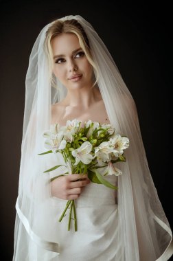 Gelin portresi, duvak ile, pencerenin yanında beyaz bir elbise içinde bir buket zambak ile. Minimalist iç mekan, karanlık arka plan, doğal aydınlatma. Makyajlı ve uzun saçlı gelin.,.