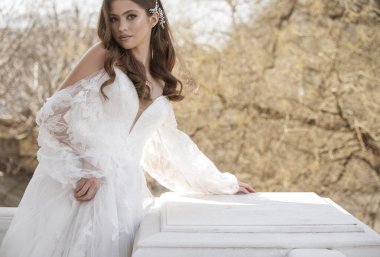 Geleneksel uzun elbiseli esmer gelin baharın başında beyaz otel balkonunda poz veriyor. Uzun kollu beyaz elbise, korse, açık omuzlar. Gelinin portresi, düğün makyajı. 