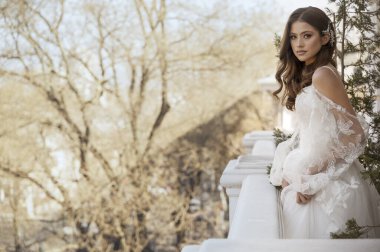 Geleneksel uzun elbiseli esmer gelin baharın başında beyaz otel balkonunda poz veriyor. Uzun kollu beyaz elbise, korse, açık omuzlar. düğün makyajı, düğün saç stili