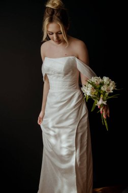 Elinde bir buket beyaz zambak olan genç sarışın gelin. Gelinin akşam makyajı ve yüksek bir saç stili var. O, karanlık bir arka planın üzerinde duruyor. Şık bir gelin ve güzellik kavramı