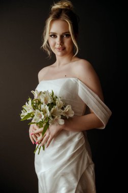 Elinde bir buket beyaz zambak olan sarışın gelin. Hayalperest gelinin akşam makyajı ve yüksek bir saç stili var. O, karanlık bir arka planın üzerinde duruyor. Zarif, açık omuzlu, şık bir elbise.