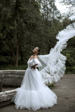 Ağaçların ve eski çeşmelerin arka planında uzun peçeli güzel bir gelin. Uzun beyaz tüylü elbiseli esmer kız gülümsüyor ve seviniyor. Rüzgarda savrulan peçeler 
