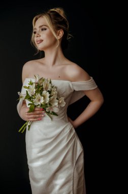 Elinde zambak buketi olan genç sarışın gelin. Gelinin akşam makyajı ve yüksek bir saç stili var. O, karanlık bir arka planın üzerinde duruyor. Açık omuzlu zarif bir elbise..