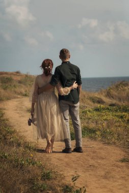 Genç aşık bir çift denize giden yolda yürüyor. Deniz kıyısında yürüyen yeni evliler. Sarılırlar, arkadan bakarlar. Açık bej elbiseli kız, sarılıyorlar. dikey yönelimin fotoğrafı