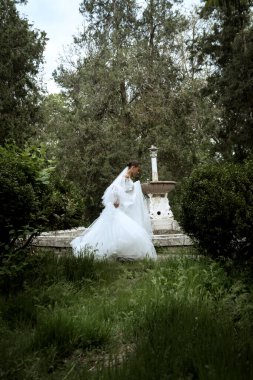 Ağaçların ve eski çeşmelerin arka planında uzun peçeli güzel bir gelin. Uzun beyaz tüylü elbiseli esmer kız gülümsüyor ve seviniyor. Peçe rüzgarda dalgalanıyor.