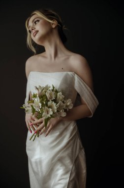 Elinde bir buket beyaz zambak olan genç sarışın gelin. Hayalperest gelinin akşam makyajı ve yüksek bir saç stili var. O, karanlık bir arka planın üzerinde duruyor. Açık omuzlu zarif bir elbise.