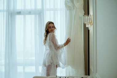 Gelinin sabahı. Dantelli cüppeli kibar ve çekici bir sarışın zarif bir gelinliğin yanında duruyor. İpek bornoz ve iç çamaşırlı genç bir kadın. Gelinin hazırlanma konsepti.